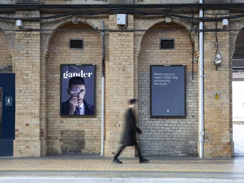A person walks past two brick wall Gander posters, one with a man holding a magnifying glass and the other with text reading "When you just need a little help uncovering the truth."